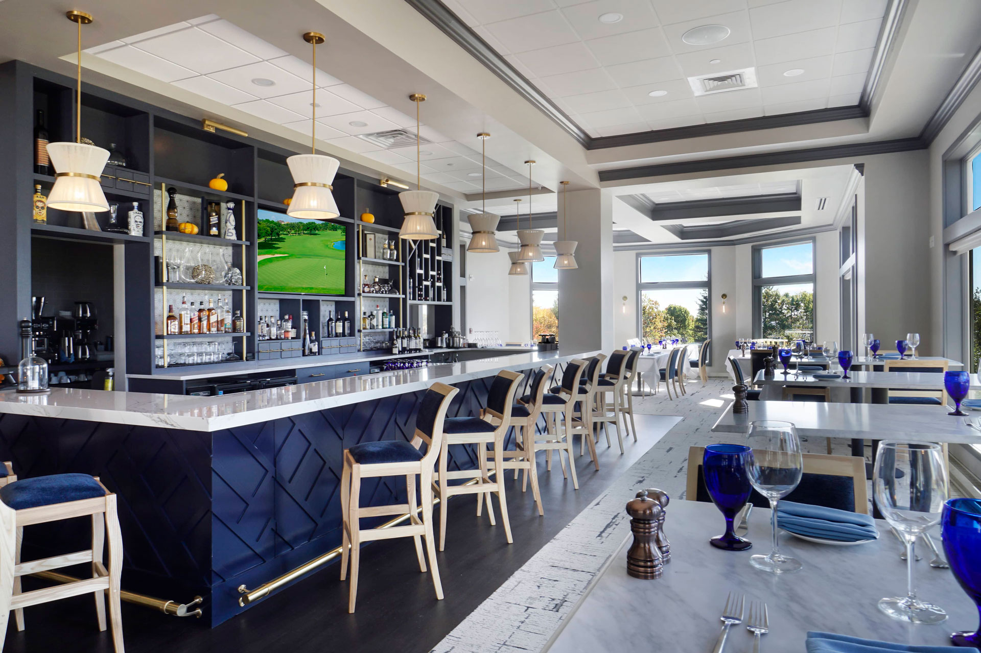 Modern restaurant interior featuring a bar with drinks, blue glassware, and a view of greenery outside.