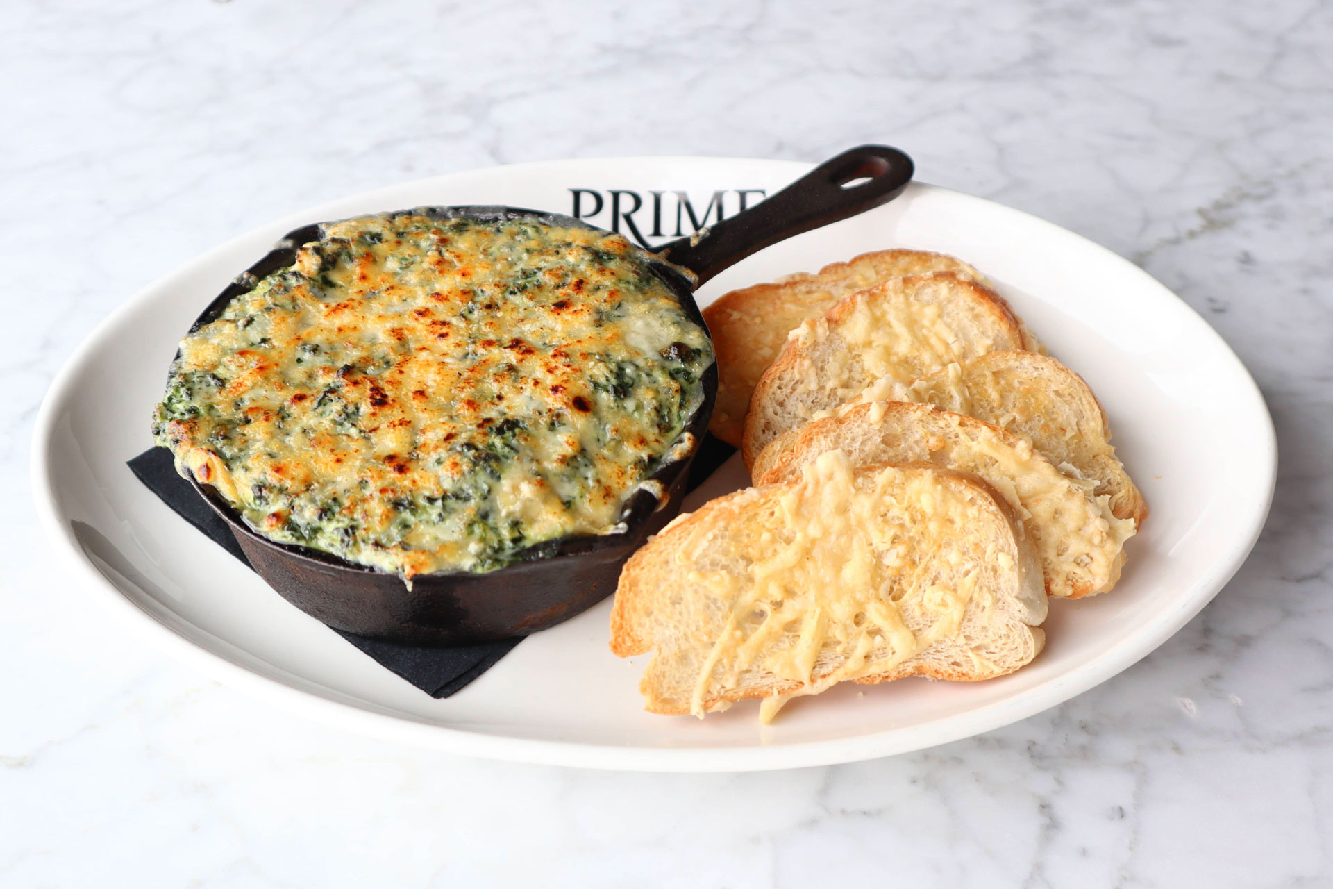 Baked spinach dip in a cast iron skillet served with toasted bread slices on a white plate.