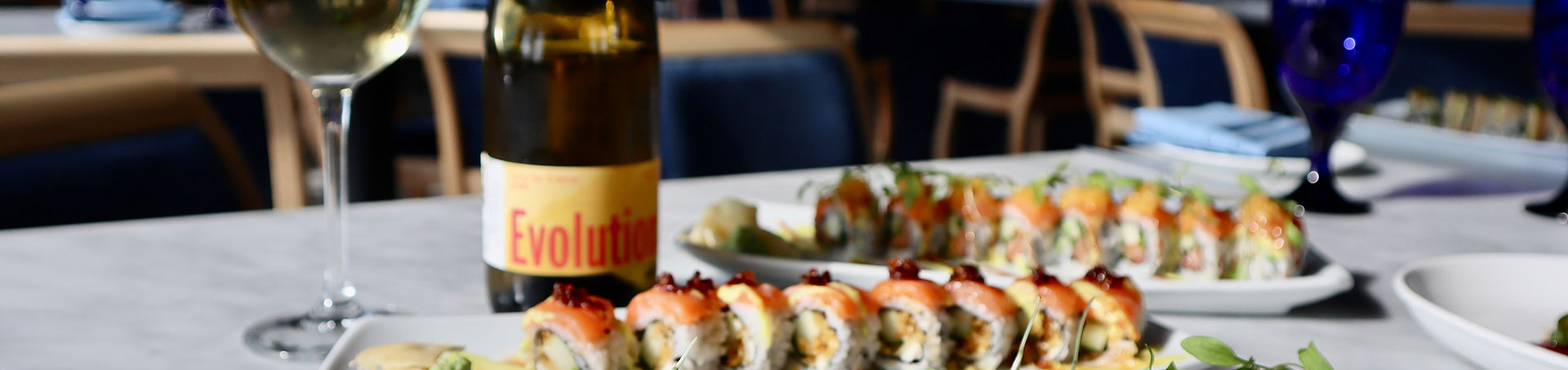 Sushi rolls and a glass of white wine on a table, with a bottle labeled 'Evolution' in the background.